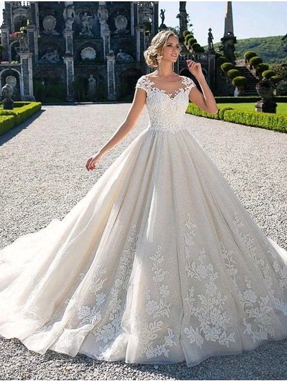 Full view of an elegant ball gown wedding dress with a jewel neck, cap sleeves, lace appliques, and a chapel train.